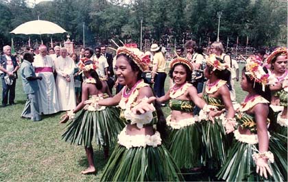Young women in bikinis and palm skirts dance in front of John Paul II